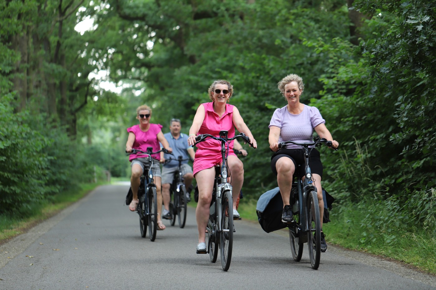 Twee vrouwen fietsen in zomerse kleding over een asfaltweg in een bosrijke omgeving. Achter hen rijden nog twee fietsers.