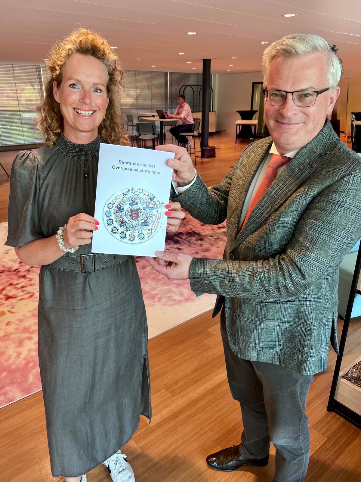 Beide personen houden het visiedocument samen omhoog. Ze staan ergens binnen met een vrij laag wit plafond. Op de grond ligt een vloerkleed. Op de achtergrond is iemand aan het werk op een laptop.