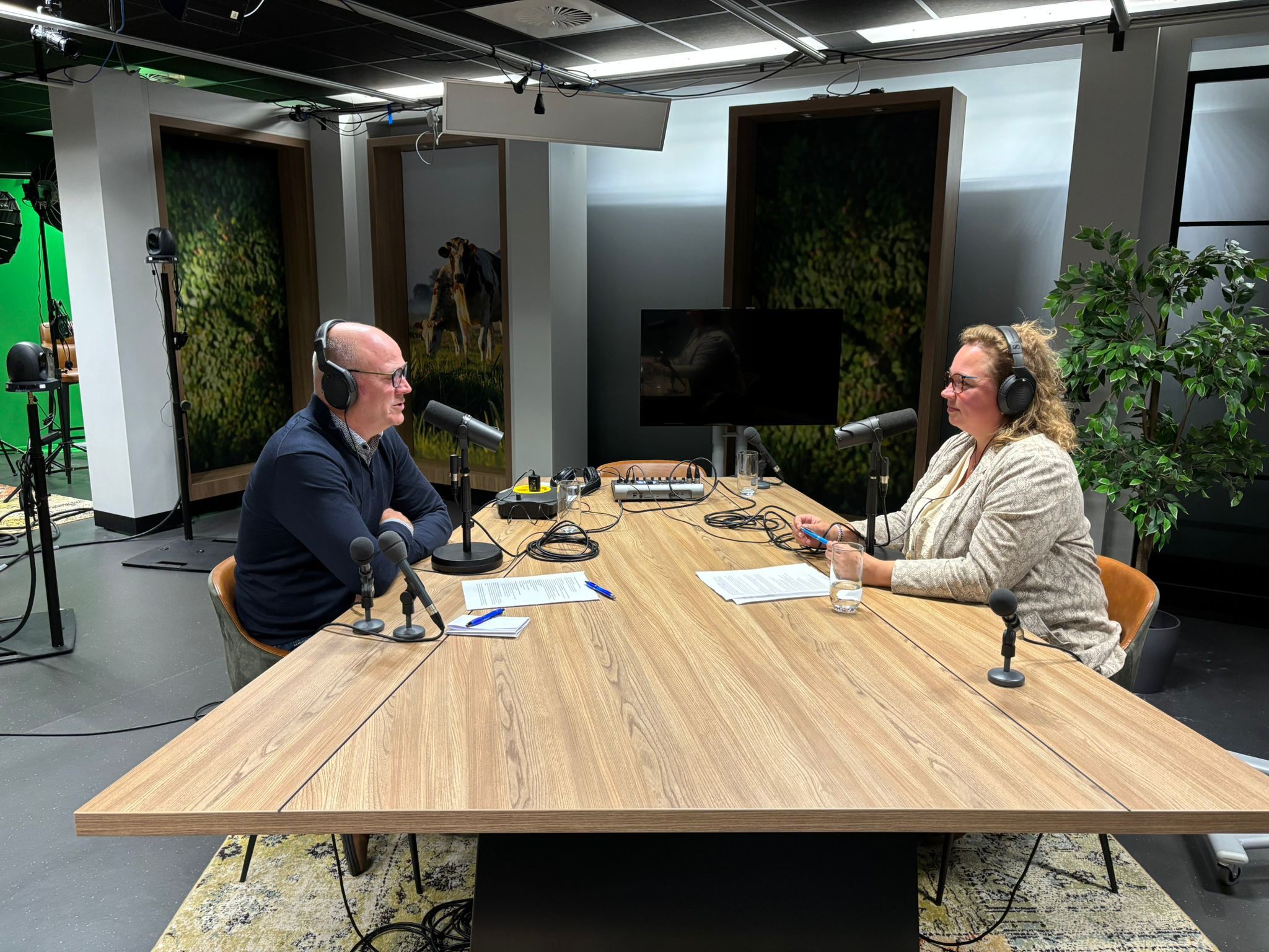 Podcaststudio met twee personen aan een houten tafel, uitgerust met microfoons, koptelefoons en opnameapparatuur. 