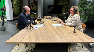 Podcaststudio met twee personen aan een houten tafel, uitgerust met microfoons, koptelefoons en opnameapparatuur. 