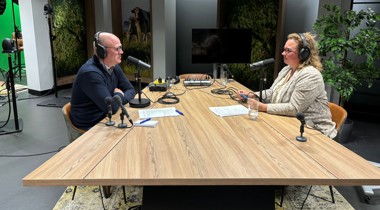 Podcaststudio met twee personen aan een houten tafel, uitgerust met microfoons, koptelefoons en opnameapparatuur. 