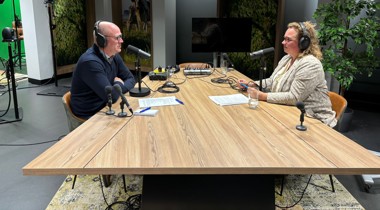 Podcaststudio met twee personen aan een houten tafel, uitgerust met microfoons, koptelefoons en opnameapparatuur. 