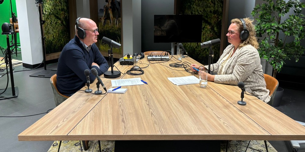 Podcaststudio met twee personen aan een houten tafel, uitgerust met microfoons, koptelefoons en opnameapparatuur. 