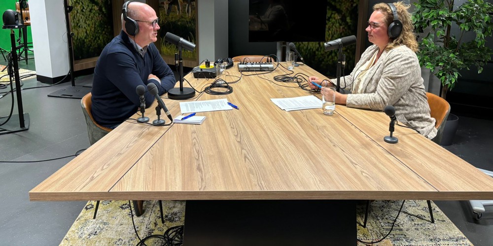 Podcaststudio met twee personen aan een houten tafel, uitgerust met microfoons, koptelefoons en opnameapparatuur. 