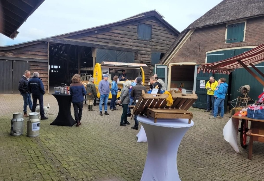 Het erf van de boerderij in Dalfsen tijdens een boer-gondische markt. Tussen de schuren staan kraampjes, bezoekers kijken rond en halen wat te eten bij een van de kraampjes. Op de voorgrond een statafel met houten kistjes waarin ook producten zijn uitgestald.