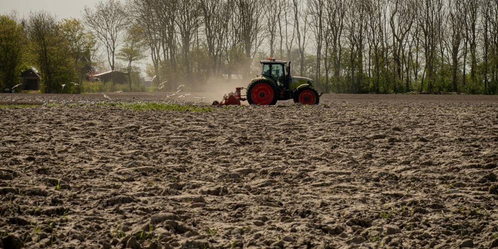 Een tractor ploegt een groot, droog veld met bomen op de achtergrond. (Foto Kenji Elzerman - Unsplash)