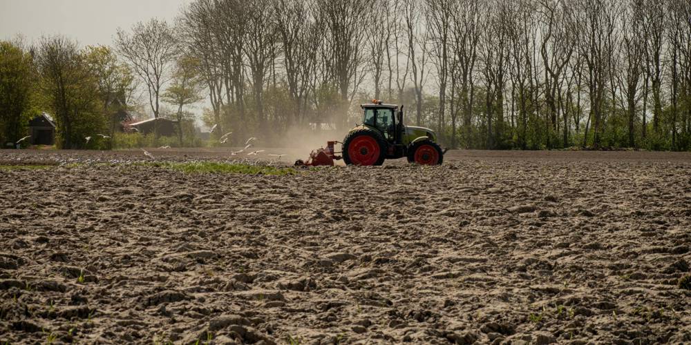 Een tractor ploegt een groot, droog veld met bomen op de achtergrond. (Foto Kenji Elzerman - Unsplash)