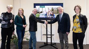 Zes mensen poseren voor een scherm terwijl twee van hen elkaar de hand schudden bij een presentatie over landbouw.