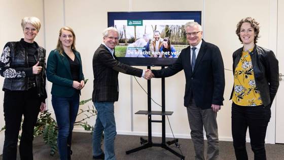 Zes mensen poseren voor een scherm terwijl twee van hen elkaar de hand schudden bij een presentatie over landbouw.