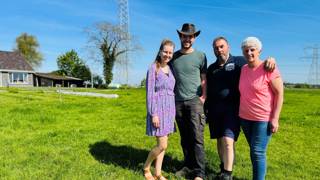Vier mensen op een grasveld met een huis, bomen, hoogspanningslijnen en een zendmast op de achtergrond. 