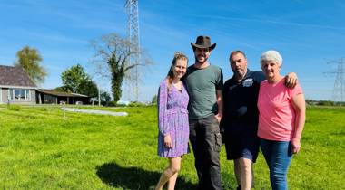 Vier mensen op een grasveld met een huis, bomen, hoogspanningslijnen en een zendmast op de achtergrond. 