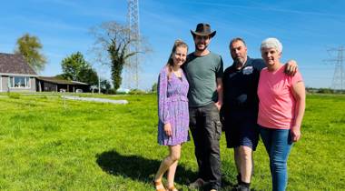 Vier mensen op een grasveld met een huis, bomen, hoogspanningslijnen en een zendmast op de achtergrond. 