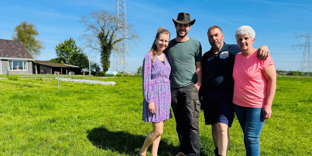 Vier mensen op een grasveld met een huis, bomen, hoogspanningslijnen en een zendmast op de achtergrond. 