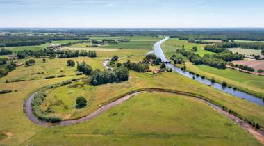 Luchtfoto van een landelijk gebied met groene velden, bomen, rivieren en verspreide huizen onder een deels bewolkte hemel.