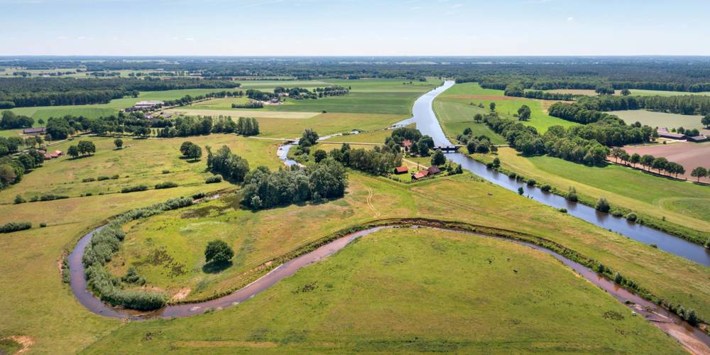 Luchtfoto van een landelijk gebied met groene velden, bomen, rivieren en verspreide huizen onder een deels bewolkte hemel.