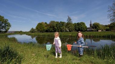 Twee jonge kinderen vangen waterdiertjes met schepnetten langs de oever van een sloot, met een kerktoren en groene bomen op de achtergrond.