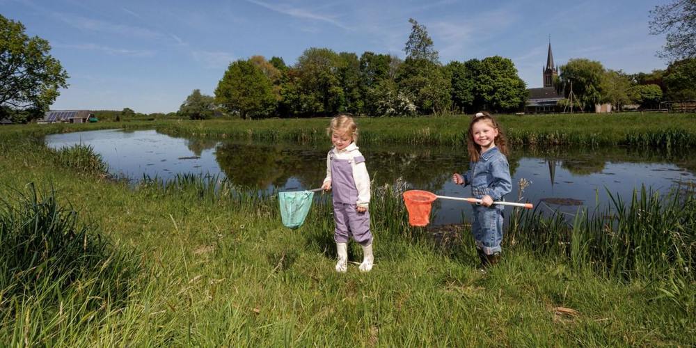 Twee jonge kinderen vangen waterdiertjes met schepnetten langs de oever van een sloot, met een kerktoren en groene bomen op de achtergrond.
