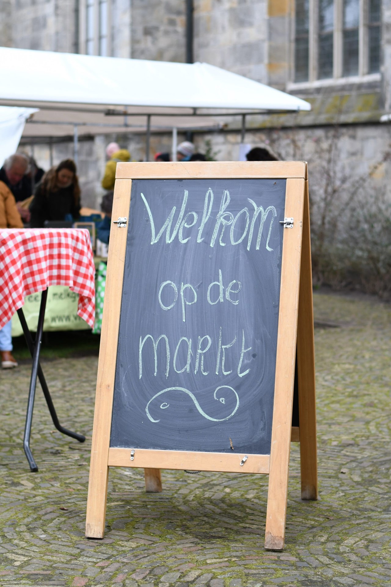 Staand kreitbord op de markt met de tekst "Welkom op de markt". Op de achtergrond de kerk bij het Kerkplein en een van de marktkramen.