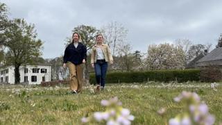 Twee vrouwen wandelen door een bloeiend grasveld met bomen en een witte boerderij op de achtergrond.