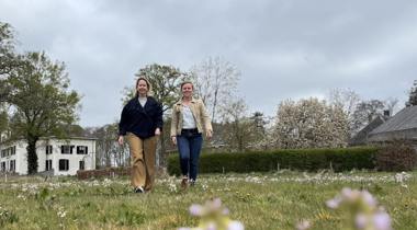 Twee vrouwen wandelen door een bloeiend grasveld met bomen en een witte boerderij op de achtergrond.