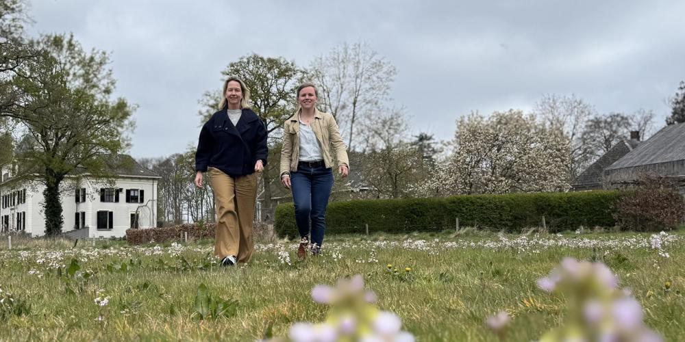 Twee vrouwen wandelen door een bloeiend grasveld met bomen en een witte boerderij op de achtergrond.