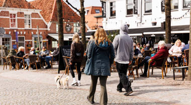 Een zonnig stadsplein met terrassen waar mensen aan een drankje zitten. Andere mensen lopen over het plein.