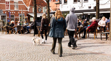 Een zonnig stadsplein met terrassen waar mensen aan een drankje zitten. Andere mensen lopen over het plein.