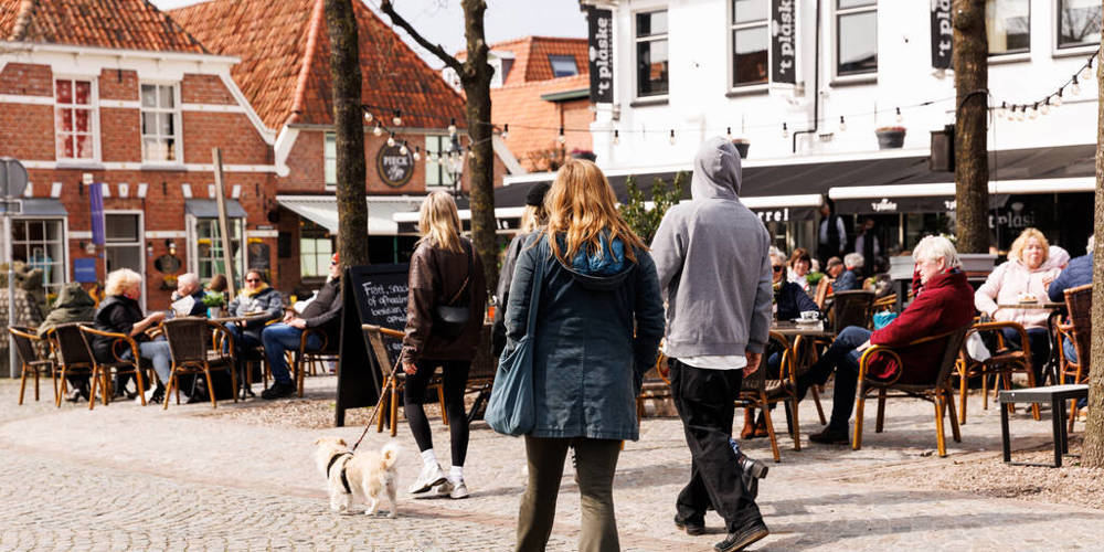 Een zonnig stadsplein met terrassen waar mensen aan een drankje zitten. Andere mensen lopen over het plein.