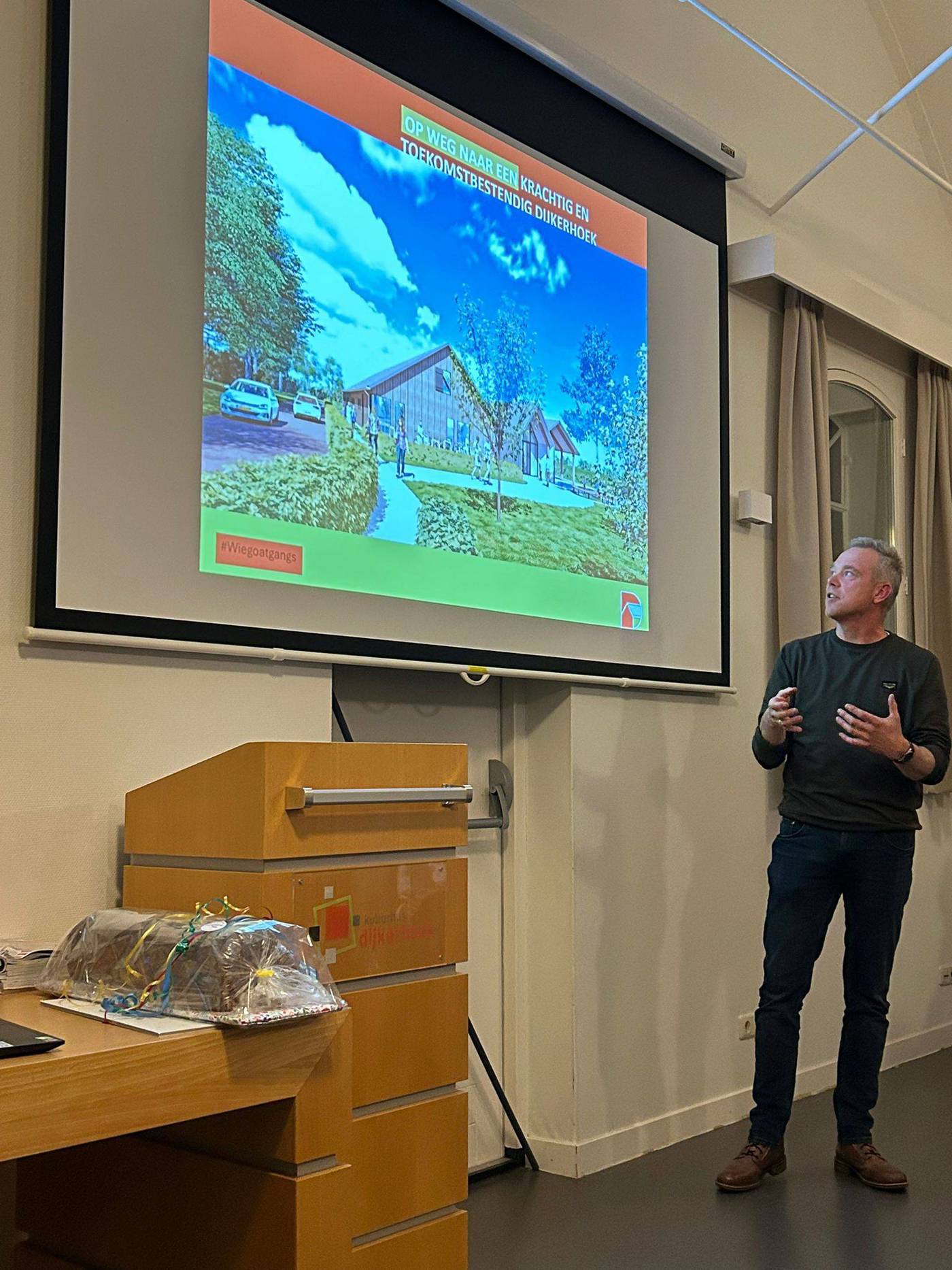 Een man geeft een presentatie met een dia waarop een visueel van een modern bouwproject wordt getoond, terwijl een ingepakt cadeau op een tafel in de voorgrond staat.