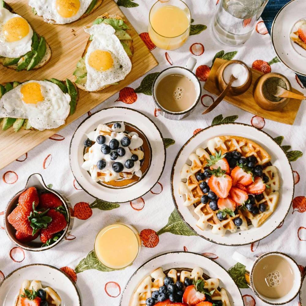 Ontbijttafel met wafels, avocado-toast met ei, aardbeien, koffie, sinaasappelsap en een koperen ketel op een fruitig tafelkleed.