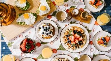 Ontbijttafel met wafels, avocado-toast met ei, aardbeien, koffie, sinaasappelsap en een koperen ketel op een fruitig tafelkleed.
