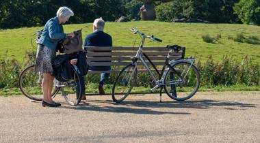 Twee fietsers nemen pauze op een bankje bij een hunebed, met fietsen en wegwijzerbord in beeld.