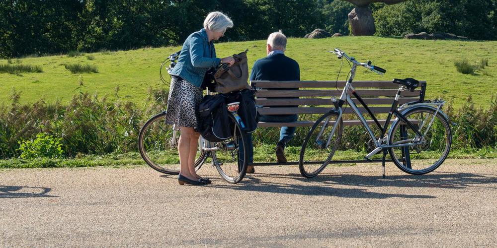 Twee fietsers nemen pauze op een bankje bij een hunebed, met fietsen en wegwijzerbord in beeld.