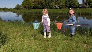 Twee jonge kinderen vangen waterdiertjes met schepnetten langs de oever van een sloot, met een kerktoren en groene bomen op de achtergrond.