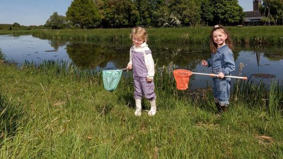 Twee jonge kinderen vangen waterdiertjes met schepnetten langs de oever van een sloot, met een kerktoren en groene bomen op de achtergrond.