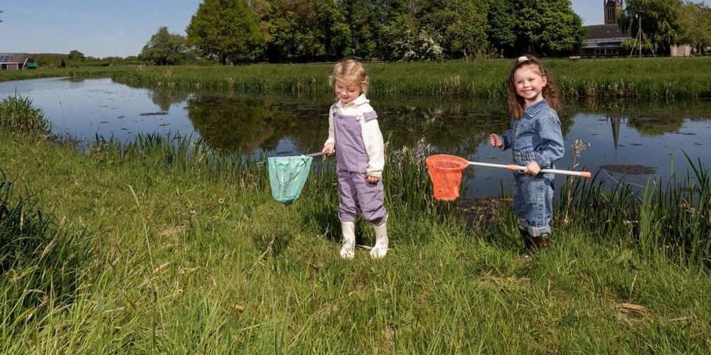 Twee jonge kinderen vangen waterdiertjes met schepnetten langs de oever van een sloot, met een kerktoren en groene bomen op de achtergrond.