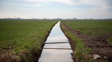 Sloot in grasland met houten plankbrug en hoogspanningslijnen in de verte onder deels bewolkte lucht.