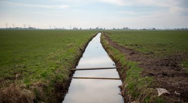 Sloot in grasland met houten plankbrug en hoogspanningslijnen in de verte onder deels bewolkte lucht.