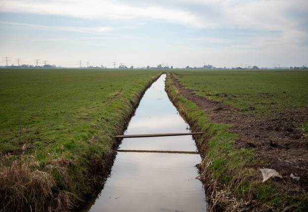 Sloot in grasland met houten plankbrug en hoogspanningslijnen in de verte onder deels bewolkte lucht.