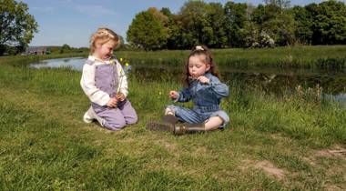 Twee kinderen zitten op het gras bij water, met bloemen en een kerk op de achtergrond.