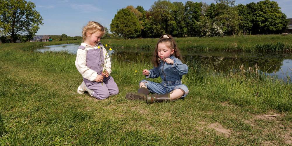 Twee kinderen zitten op het gras bij water, met bloemen en een kerk op de achtergrond.