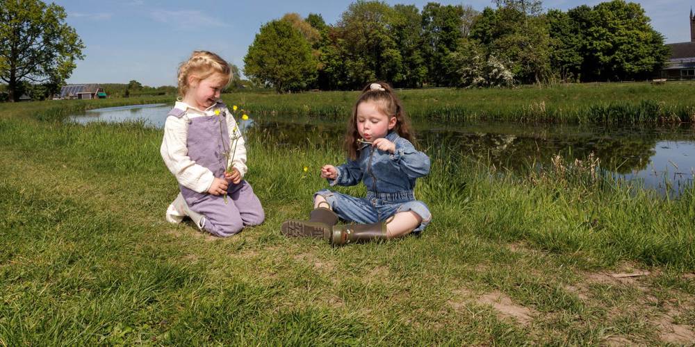 Twee kinderen zitten op het gras bij water, met bloemen en een kerk op de achtergrond.