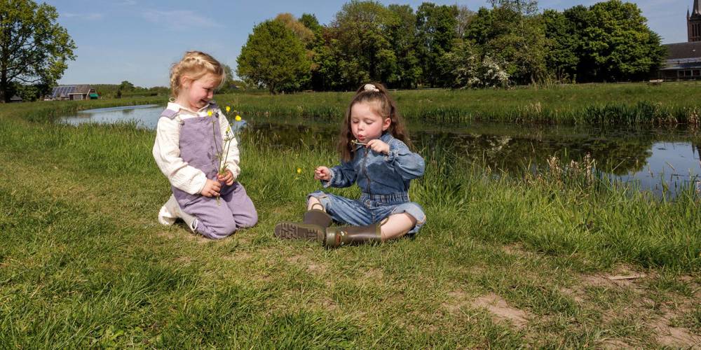 Twee kinderen zitten op het gras bij water, met bloemen en een kerk op de achtergrond.