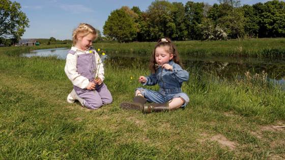 Twee kinderen zitten op het gras bij water, met bloemen en een kerk op de achtergrond.