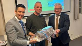 Drie mensen staan samen, waarbij één een ingepakt cadeau vasthoudt, terwijl ze glimlachen tijdens een presentatie in een vergaderruimte.