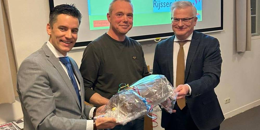 Drie mensen staan samen, waarbij één een ingepakt cadeau vasthoudt, terwijl ze glimlachen tijdens een presentatie in een vergaderruimte.