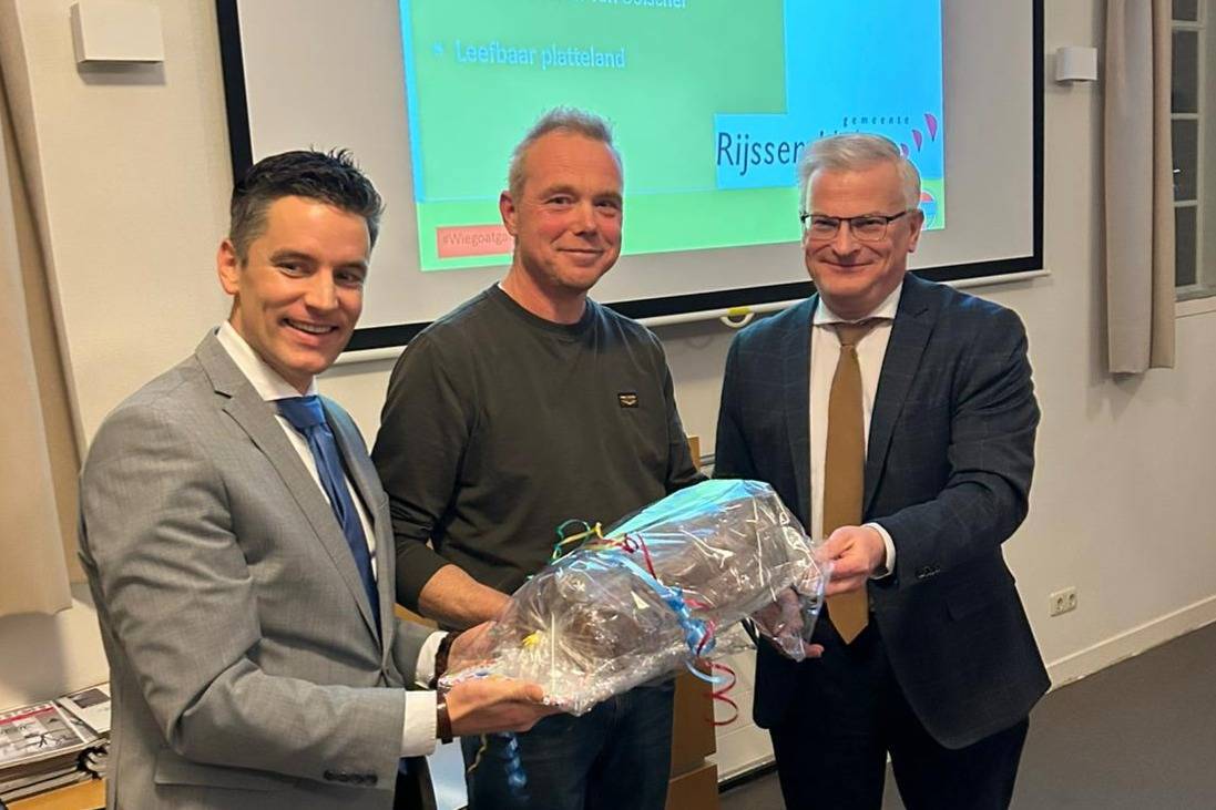 Drie mensen staan samen, waarbij één een ingepakt cadeau vasthoudt, terwijl ze glimlachen tijdens een presentatie in een vergaderruimte.