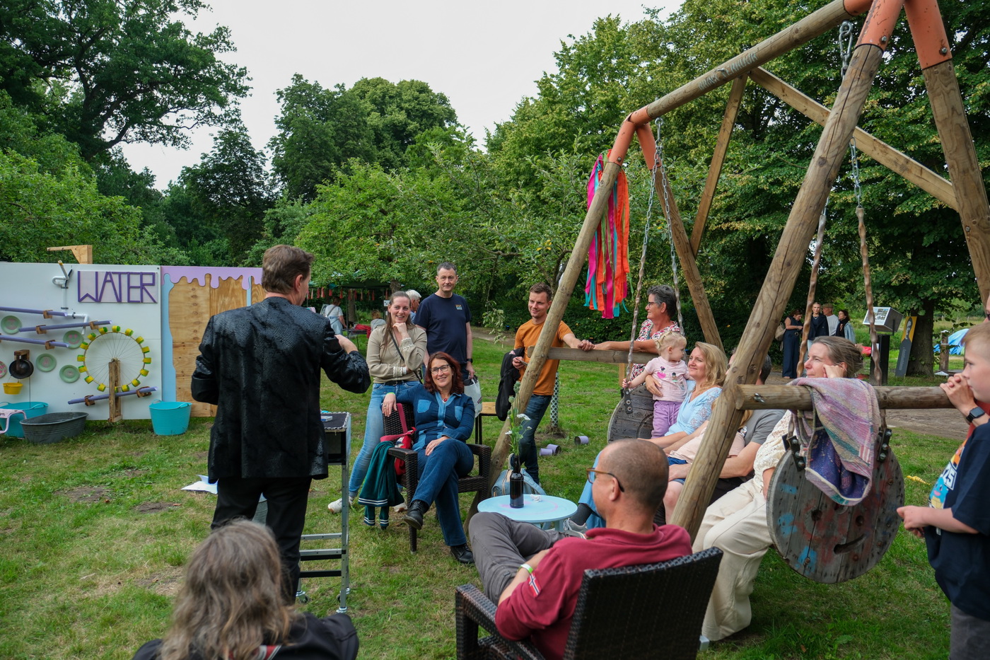 Een groepje bezoekers van het festival staan en zitten op een houten schommelbank en stoelen en luisteren naar een spreker bij een locatie op het graf van het festivalterrein. Op de achtergrond staat een wand rond het thema water. Er staan veel bomen in de omgeving en op de achtergrond lopen meer mensen.