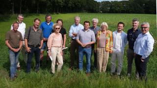 Groepsfoto van dertien mensen die buiten op een grasveld staan met een groene akker en bomen op de achtergrond, onder een blauwe lucht met wolken.