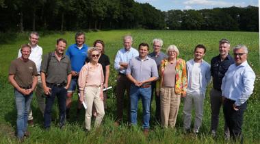 Groepsfoto van dertien mensen die buiten op een grasveld staan met een groene akker en bomen op de achtergrond, onder een blauwe lucht met wolken.